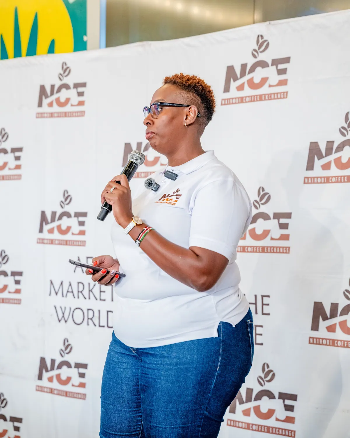 A Nairobi Coffee Exchange speaker with a microphone in front of the NCE logo wall