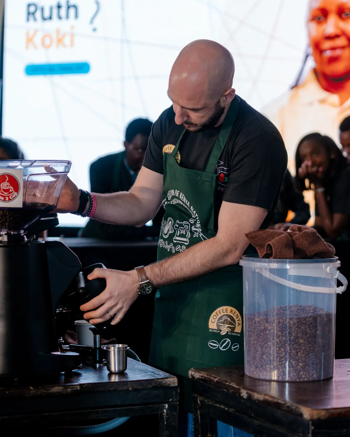 A barista in a green apron grinding coffee beside a tub of beans